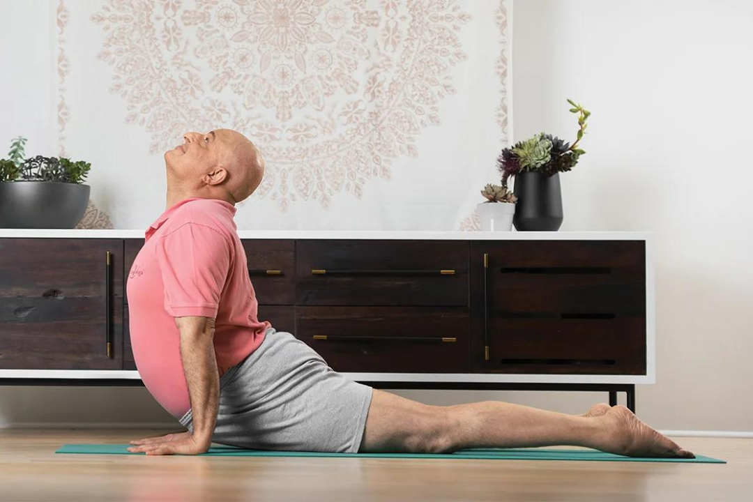 Aadil in a backbend. He is a light-skinned man without hair, wearing a pink shirt and grey shorts. The room looks serene, with a large embroidered tapestry on the wall and houseplants on the sideboard.