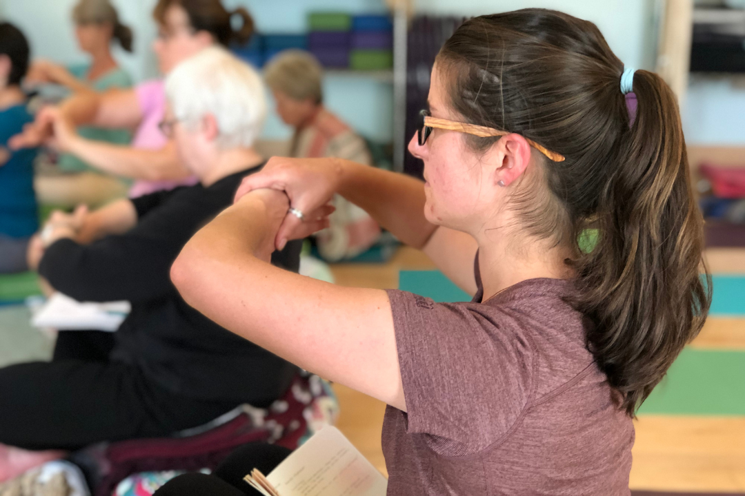 Close-up of a student in a Yoga for Healthy Hands workshop. She is wearing a dark mauve t-shirt and glasses and is sitting on the floor, holding her left wrist in her right hand out in front of her at chin level. Other students are visible in the background.
