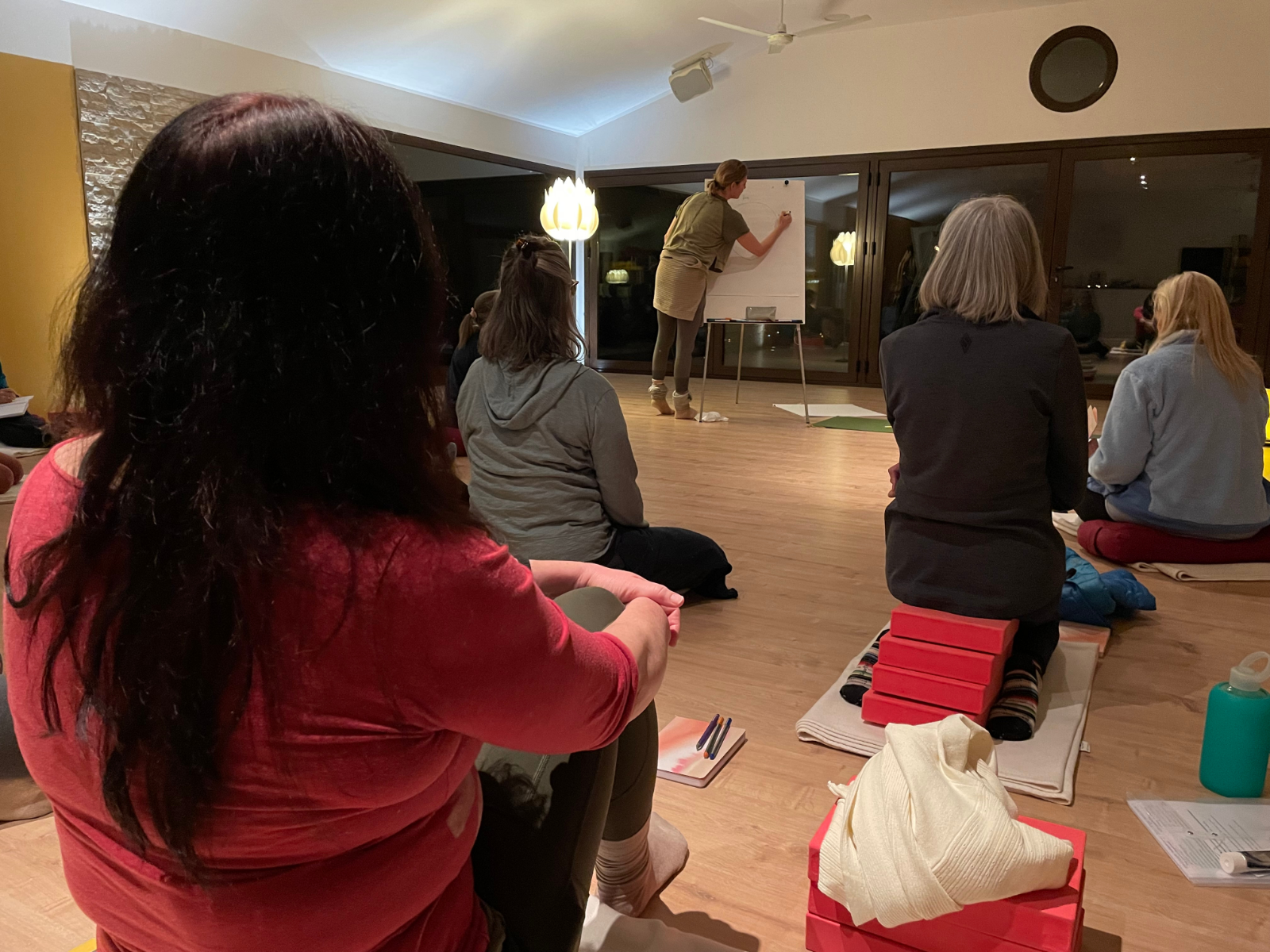 Students sit, gathered around and watch an instructor write on a white board