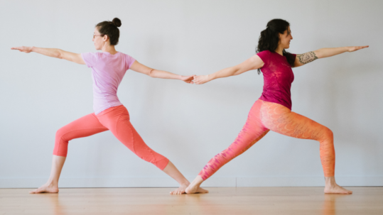 A photo of Letitia and Rebekah in Virabhadrasana II. They are facing away from each other, with their back feet touching and their back hands clasped. Their back bodies together form a vague heart shape.