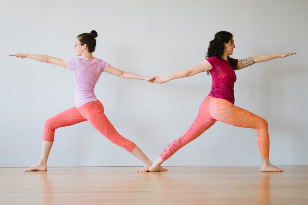 A photo of Letitia and Rebekah in Virabhadrasana II. They are facing away from each other, with their back feet touching and their back hands clasped. Their back bodies together form a vague heart shape.